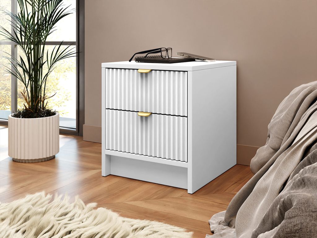 A white nightstand with two drawers and gold handles is placed next to a bed. A plant in a ribbed pot is on the left, and a fluffy rug is in the foreground.