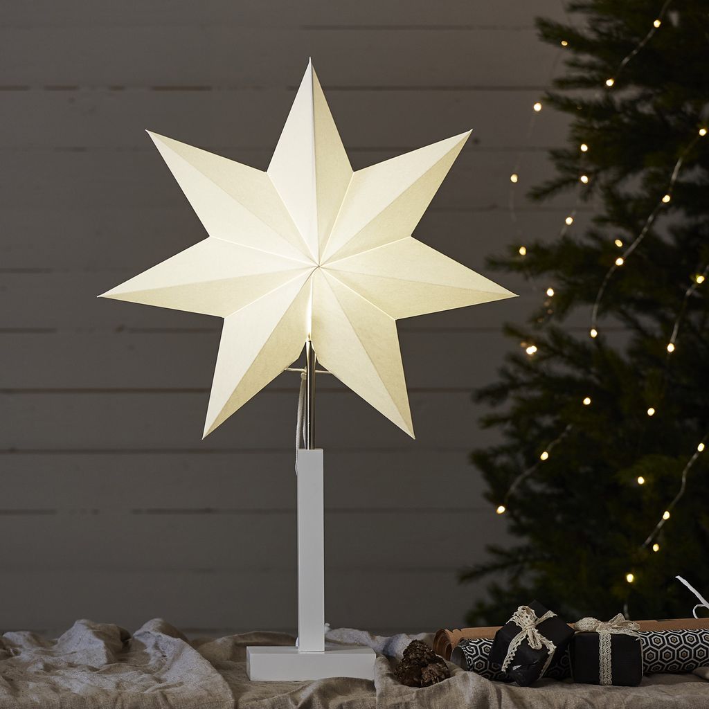 A white star-shaped lamp on a stand, placed on a table with gifts and a Christmas tree in the background.