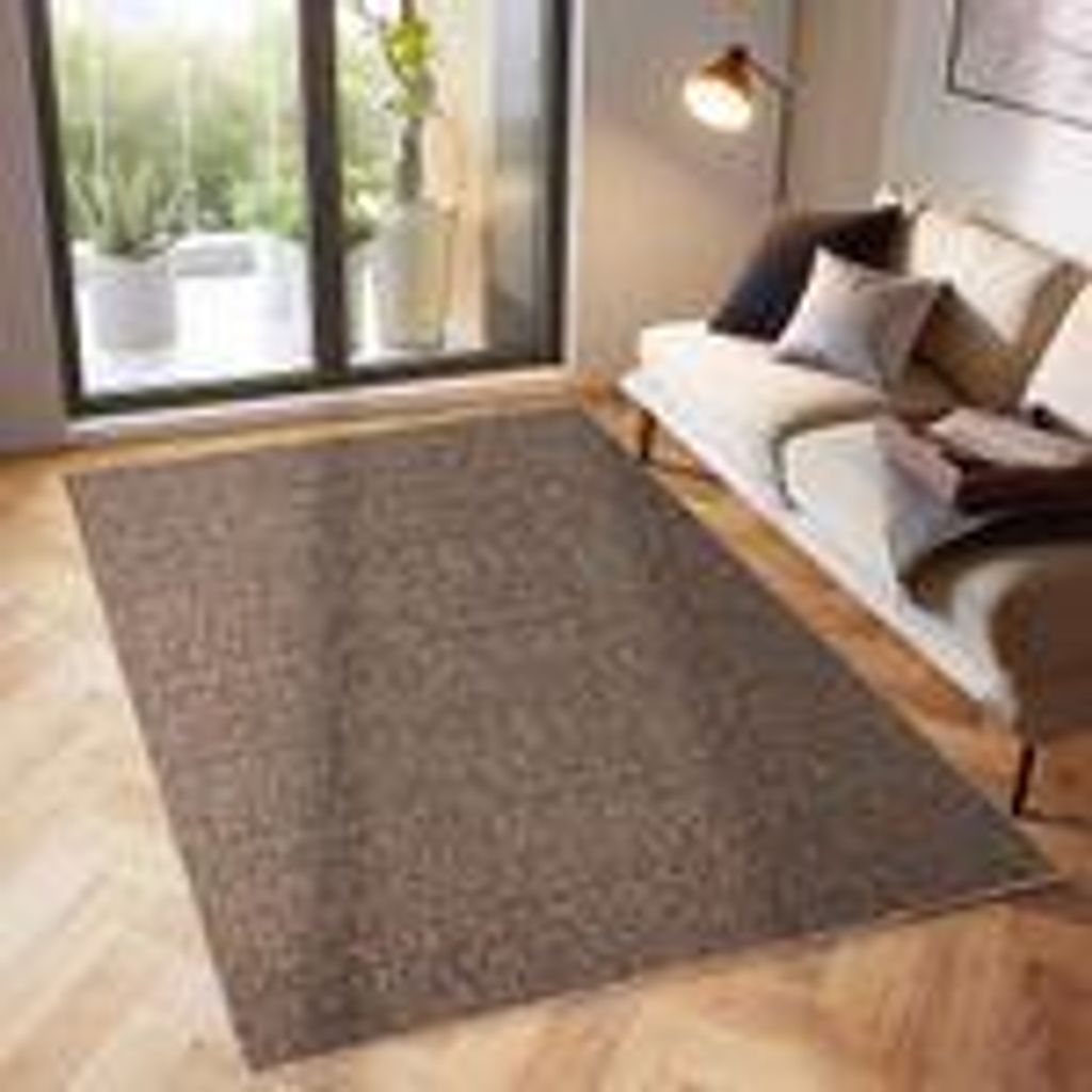 A brown rug is placed on a wooden floor in front of a sofa and a large window.