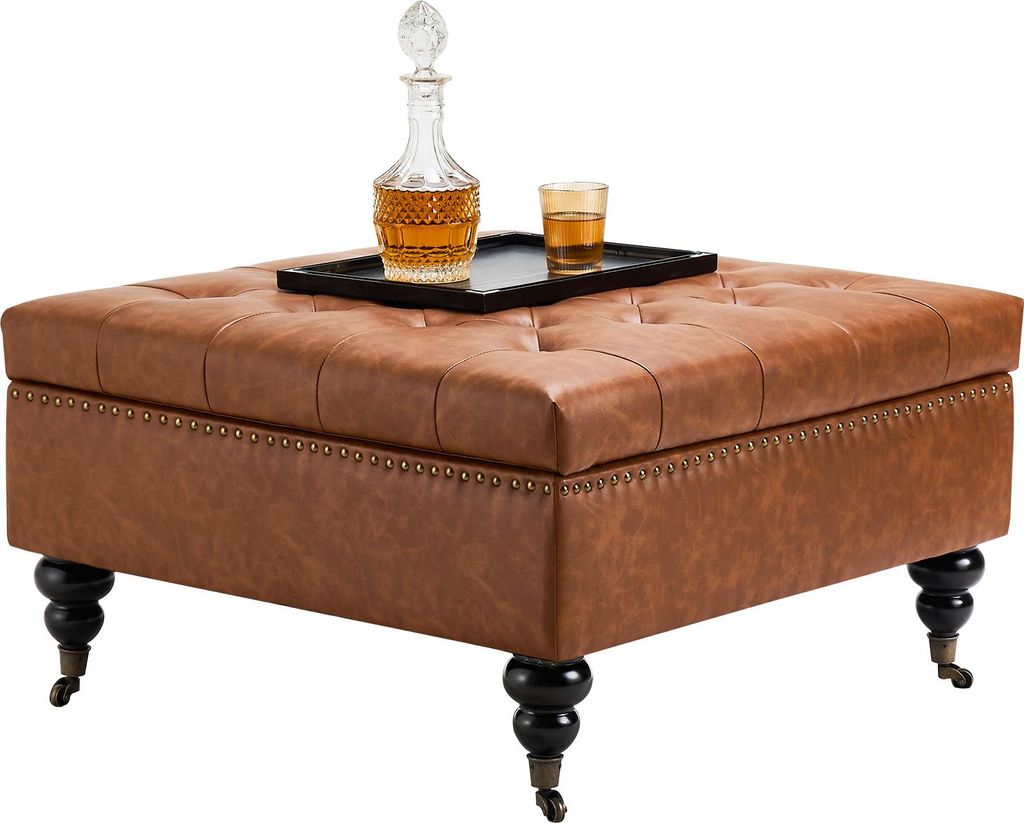 A square leather ottoman with tufted top and decorative nailhead trim sits on black legs with casters. A decanter and glass of amber liquid are on a black tray on top of the ottoman.