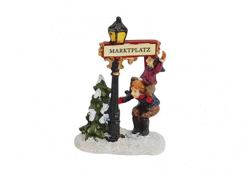 A miniature Christmas village scene with two children interacting with a lamppost and a small Christmas tree.