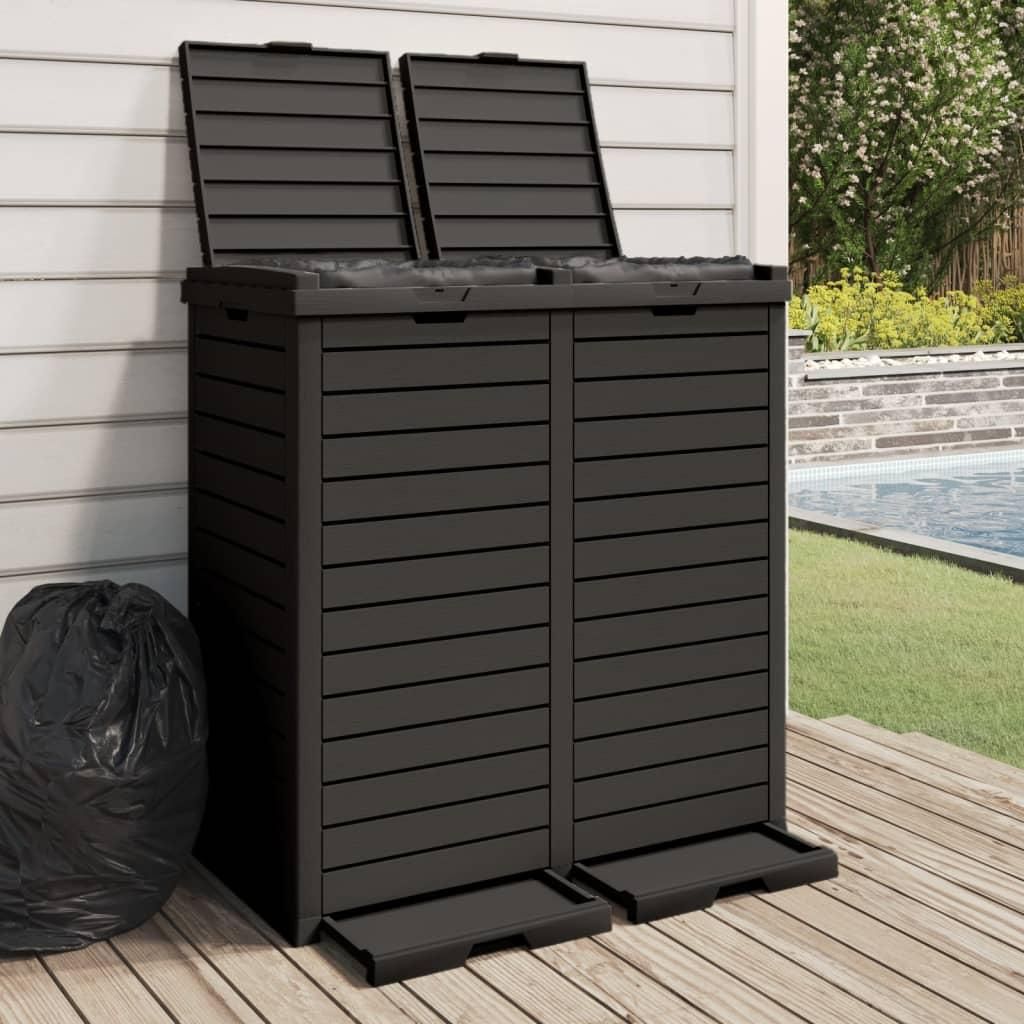 A dark gray outdoor trash can with two compartments, placed on a wooden deck next to a swimming pool.