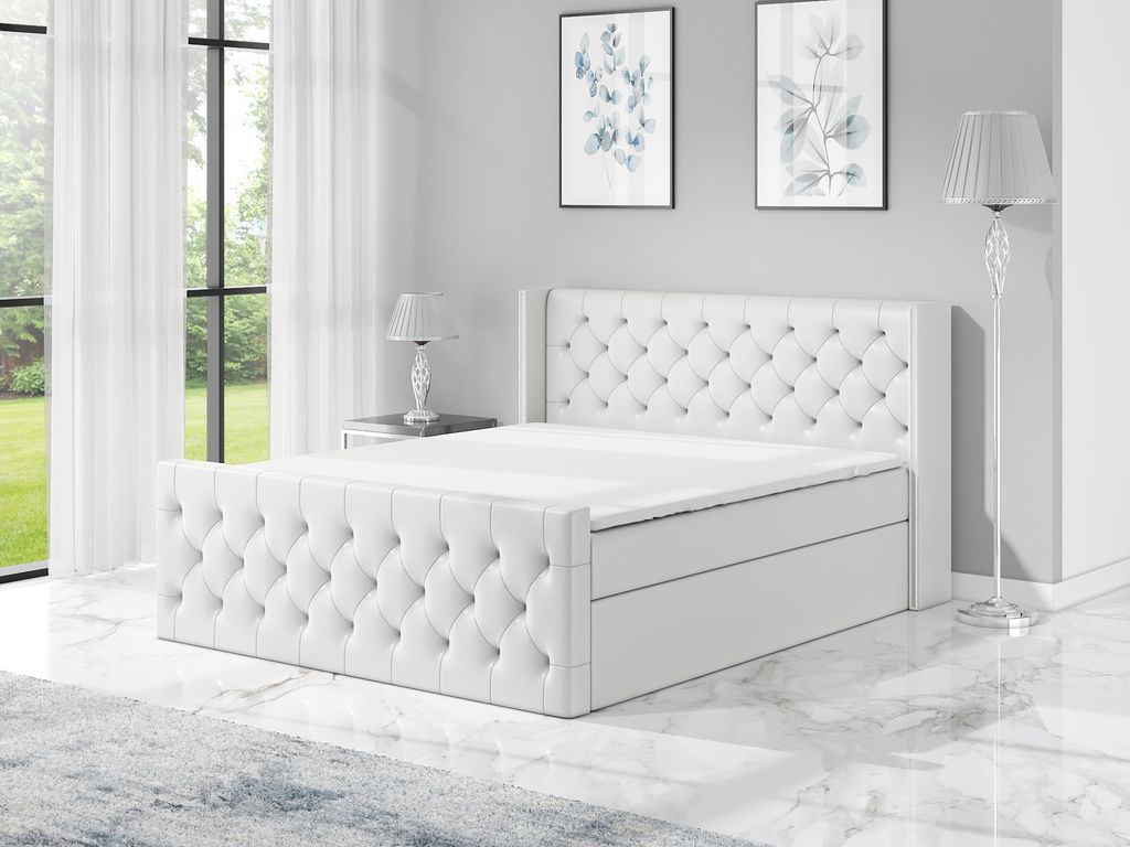 A white boxspring bed with a tufted headboard and footboard, set in a modern bedroom with large windows and artwork on the wall.