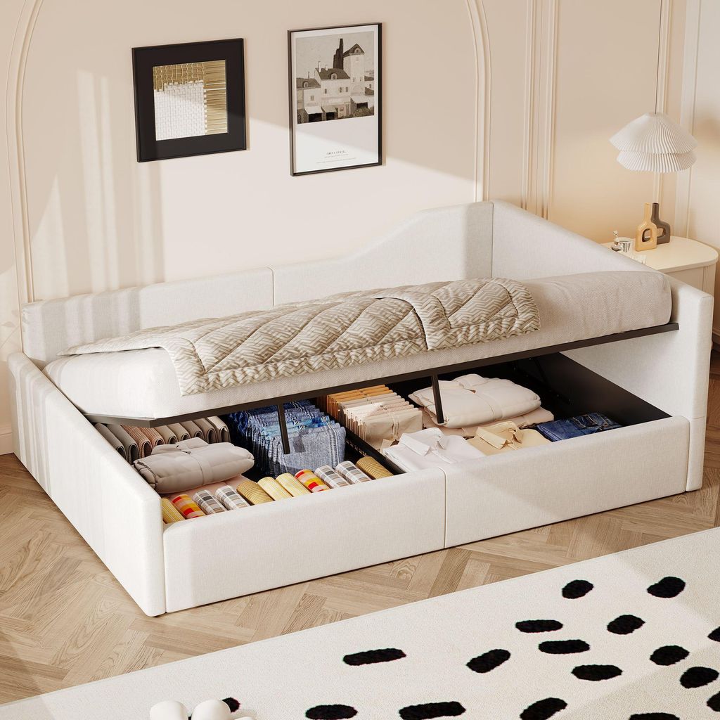 A white daybed with storage drawers is shown with the mattress lifted to reveal the storage compartments filled with folded clothes and other items. The daybed is in a room with two framed pictures on the wall and a bedside table with a lamp.