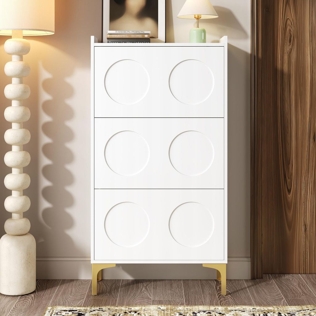 A white shoe cabinet with circular door fronts and gold legs, placed in a room with a floor lamp and a wooden door.
