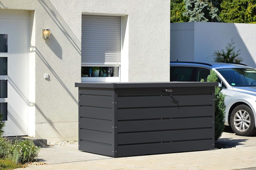 A dark gray storage box is placed on a paved area next to a white house and a silver car.