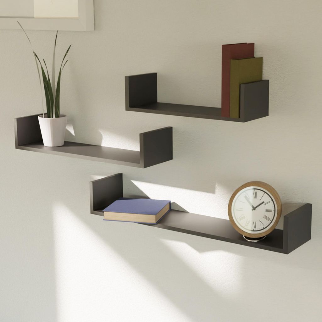 Three dark gray U-shaped shelves mounted on a light gray wall. The top shelf holds a potted plant and two books. The middle shelf holds two books. The bottom shelf holds a book and a clock.
