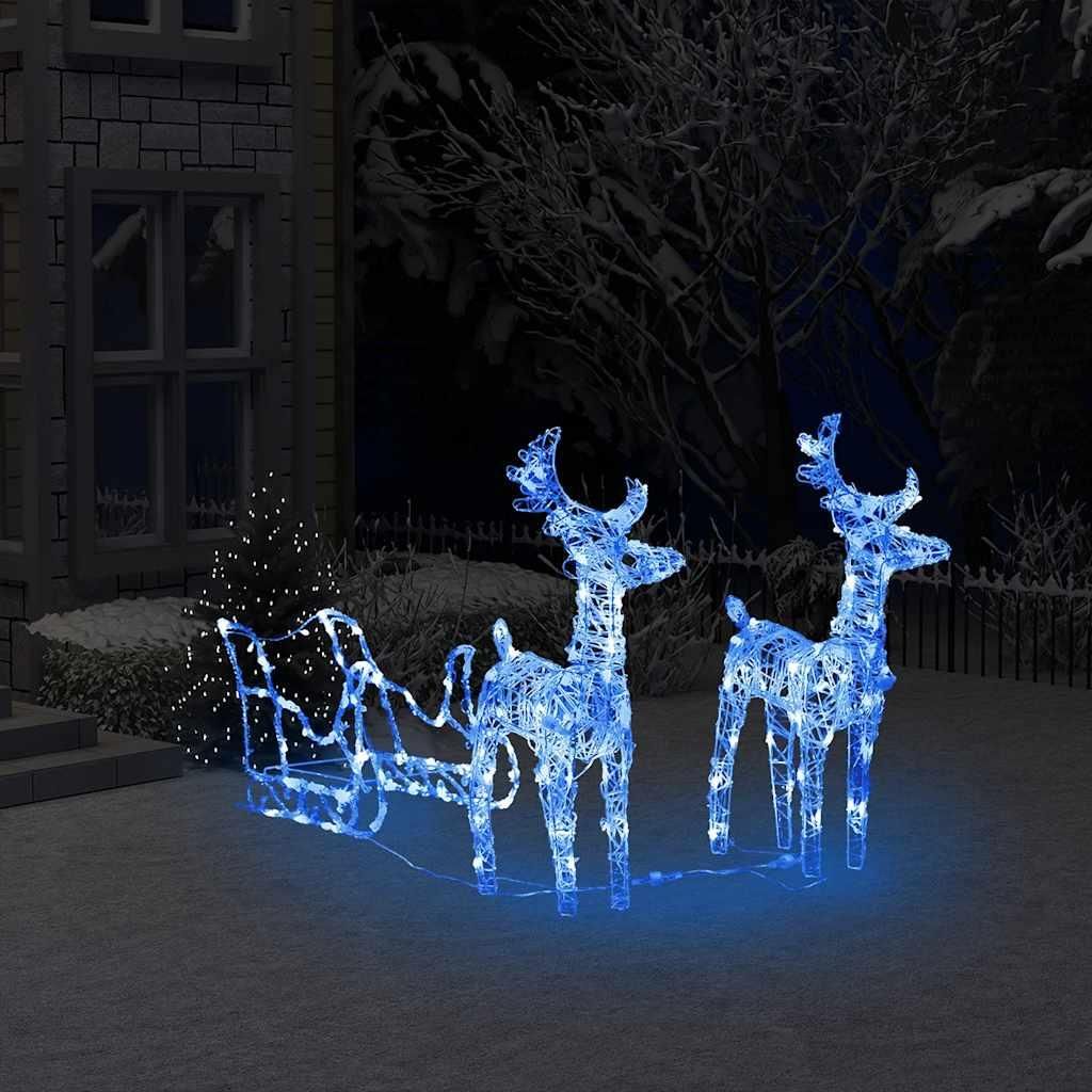 Christmas decoration of reindeer and sleigh illuminated with blue and white lights, set up in a snowy yard next to a house at night.