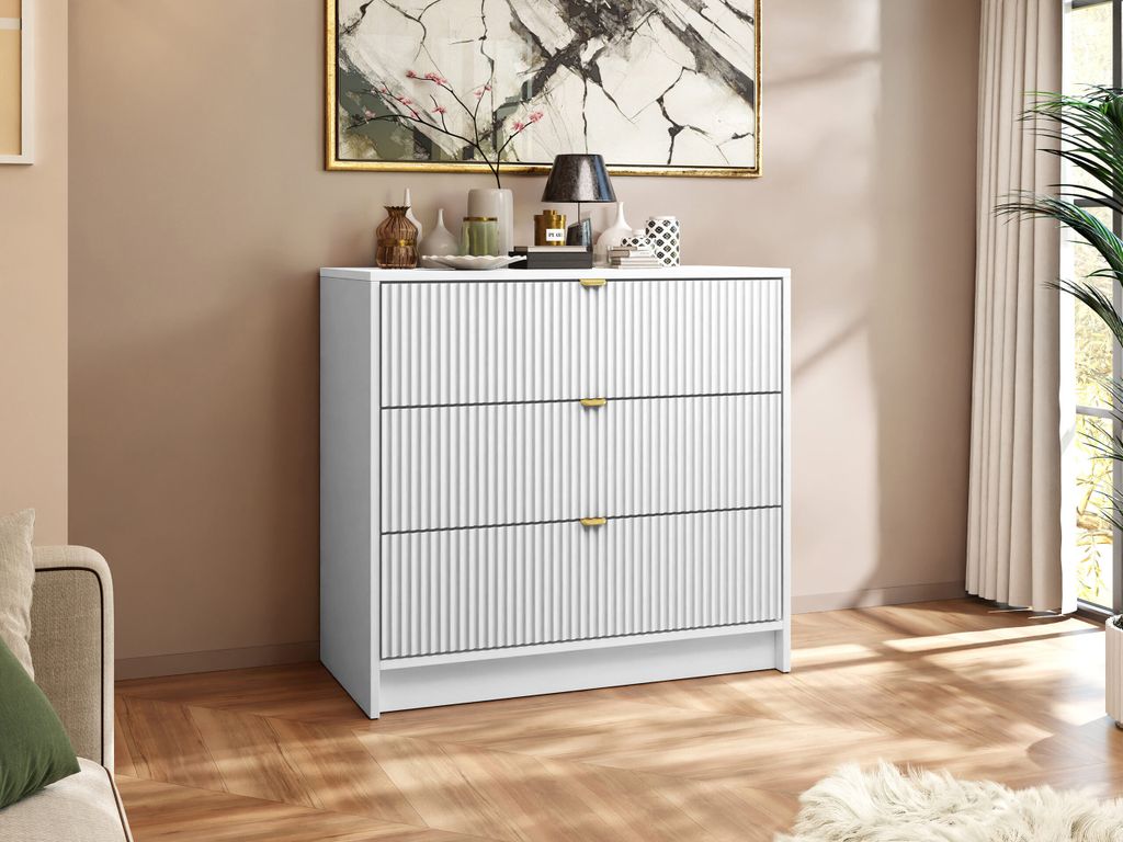 A white dresser with gold handles is shown in a room with a painting on the wall and a plant in the background.