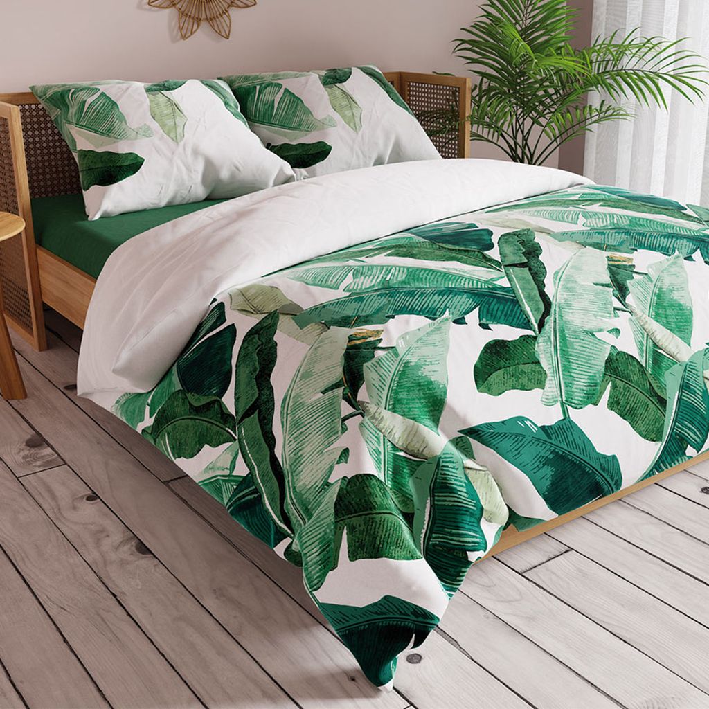 A bed with green and white banana leaf patterned bedding, set in a bedroom with wooden flooring and a plant in the background.