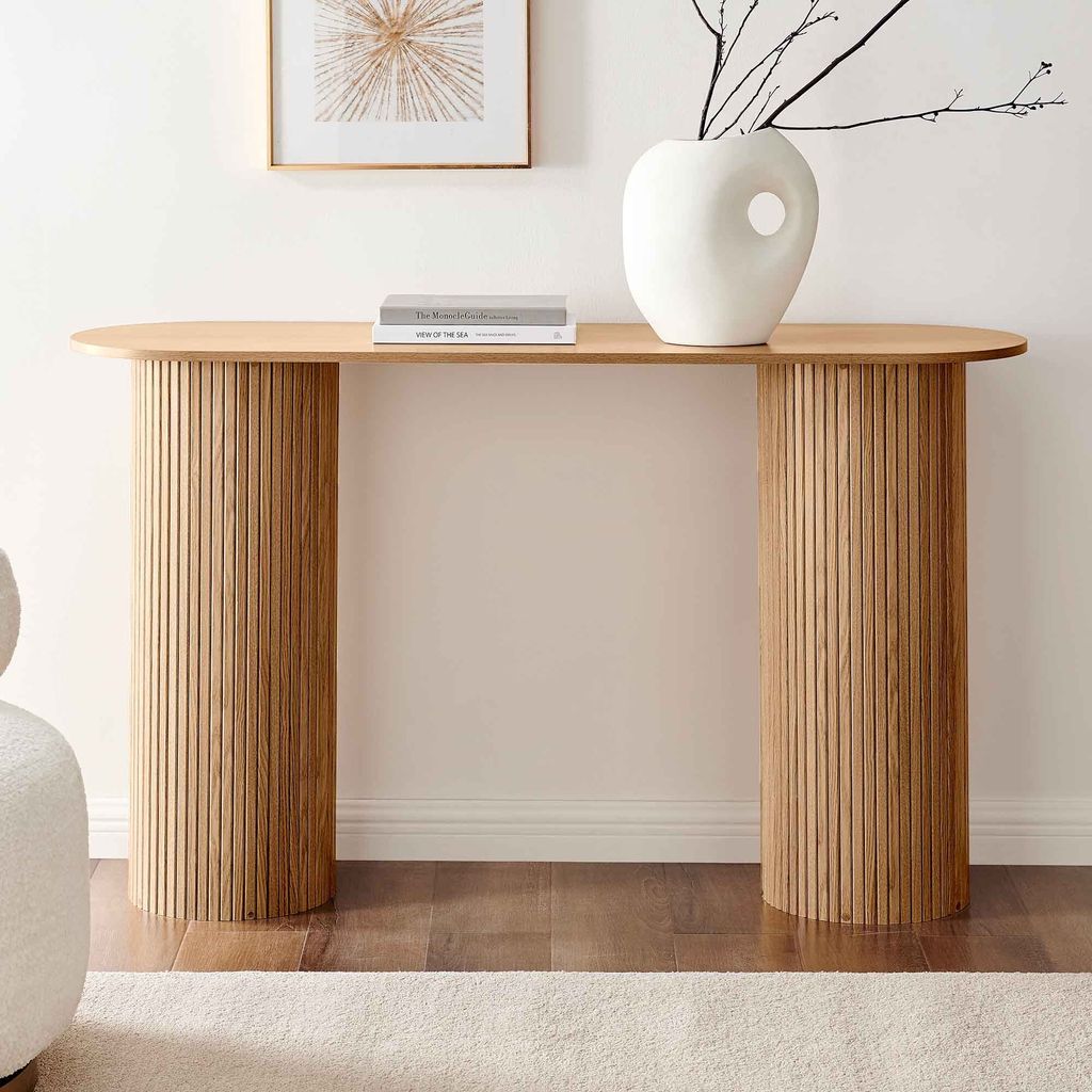 A minimalist wooden console table with a curved top and fluted cylindrical legs, placed against a white wall. A white vase with branches and two books are on the table. A portion of a white armchair and a cream rug are visible in the foreground.