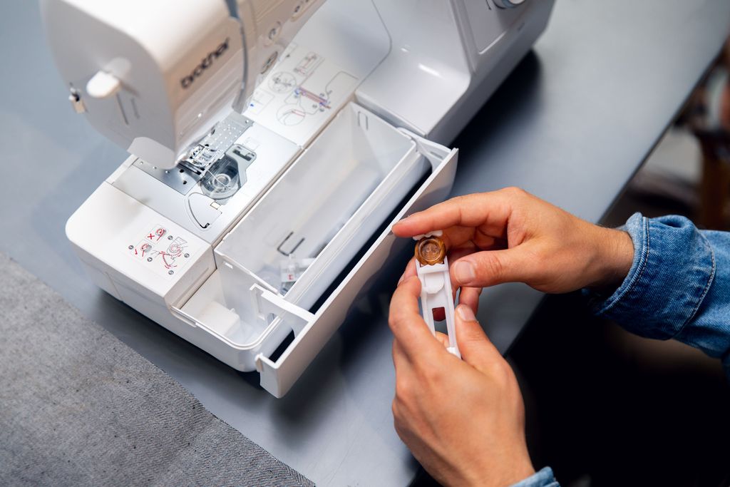 A person's hands are shown holding a bobbin and bobbin case near an open sewing machine.