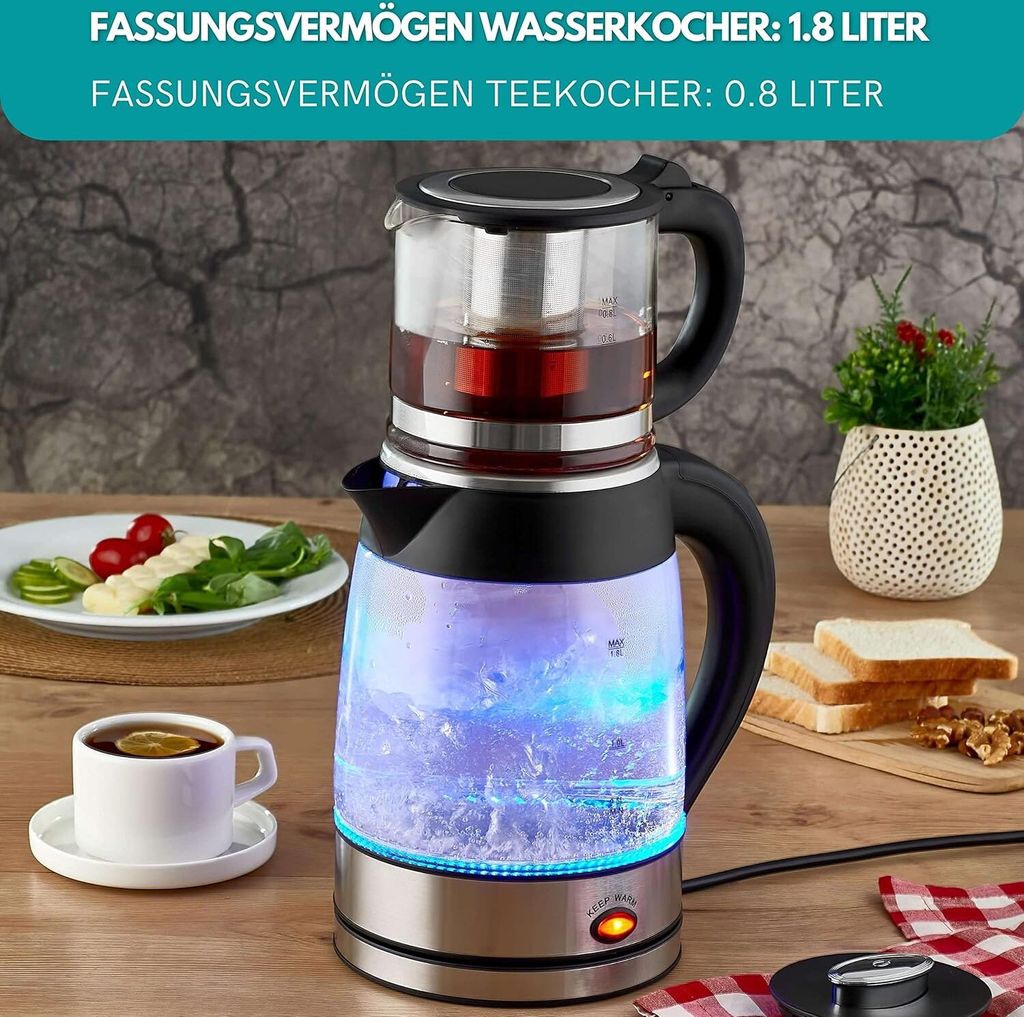 A glass electric kettle with a tea infuser on top, filled with water and illuminated by blue LEDs, sits on a wooden table next to a plate of food and a cup of tea.