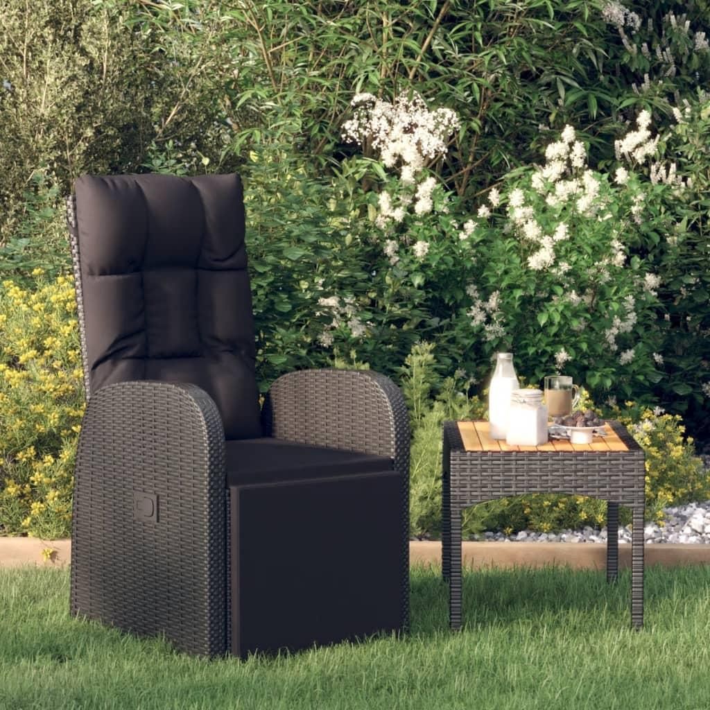 A black rattan garden recliner chair with a cushion and a small side table with a bottle and a bowl of fruit, placed on a green lawn with lush green foliage and white flowers in the background.