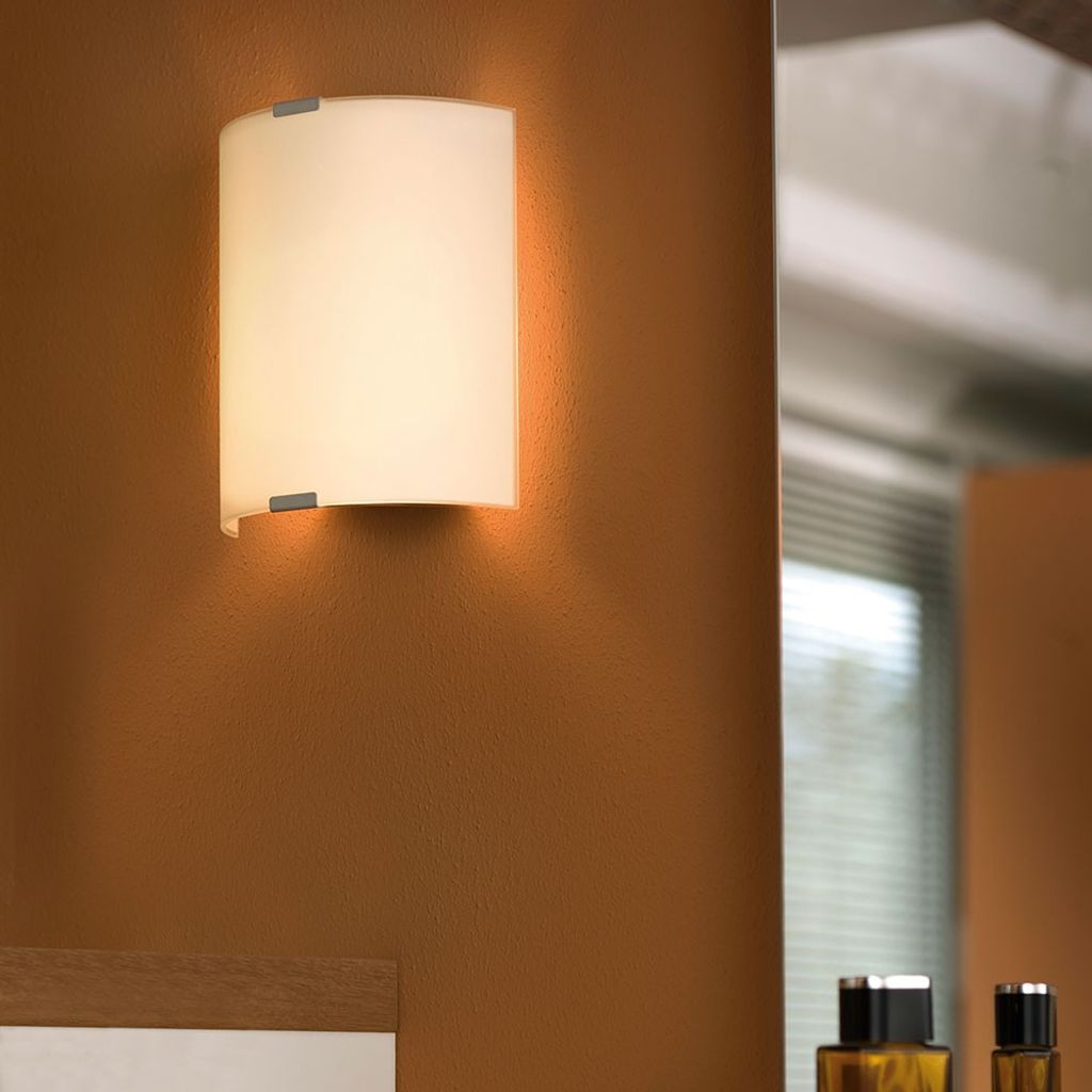 A wall-mounted light fixture with a white frosted glass shade is shown on a brown wall. A mirror is visible to the right, reflecting a window and some bottles.