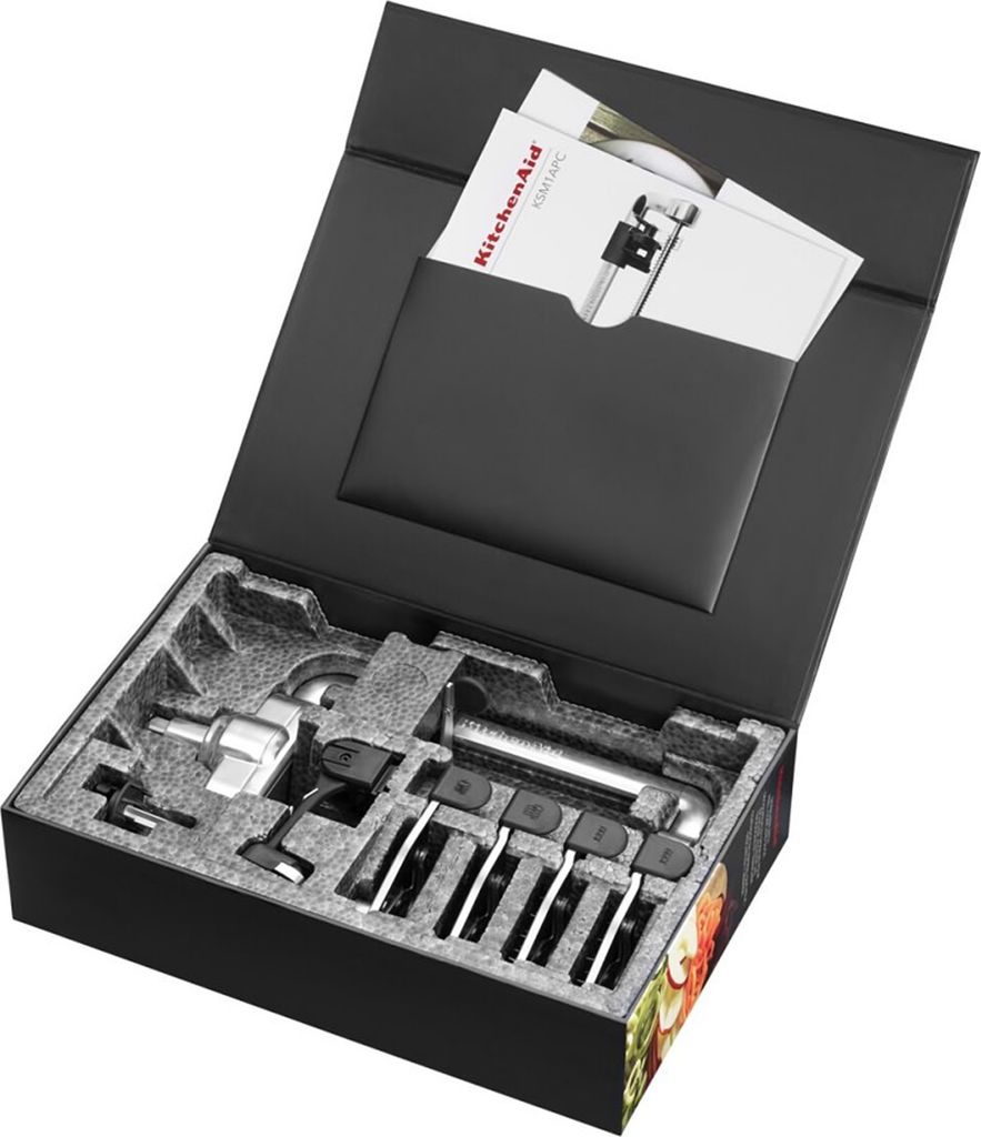 A black box opened to reveal KitchenAid food processor attachments nestled in molded foam packaging. Manuals are tucked into a black pocket on the inside lid.