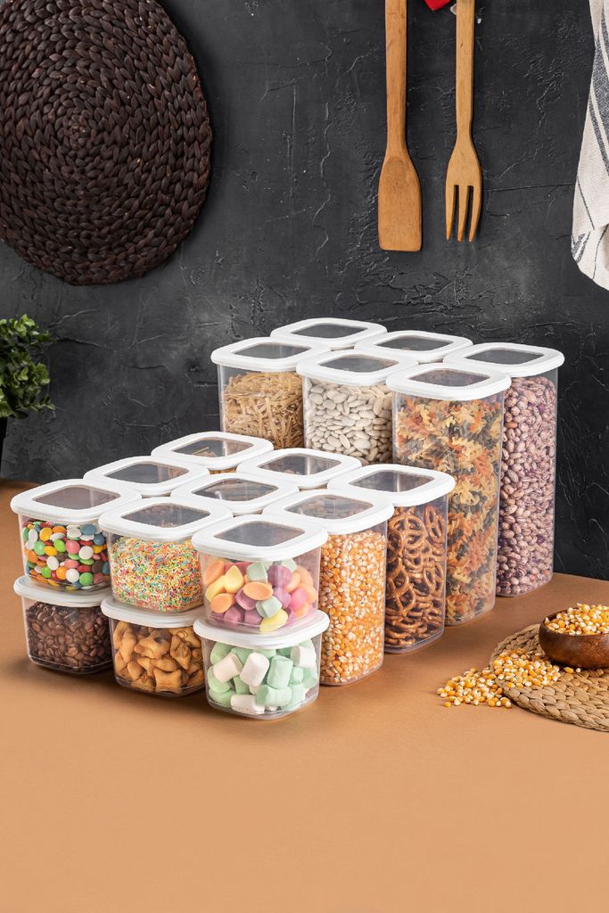 A collection of clear plastic food storage containers filled with various dry goods and candies, arranged on a brown surface against a dark textured wall with wooden utensils hanging.