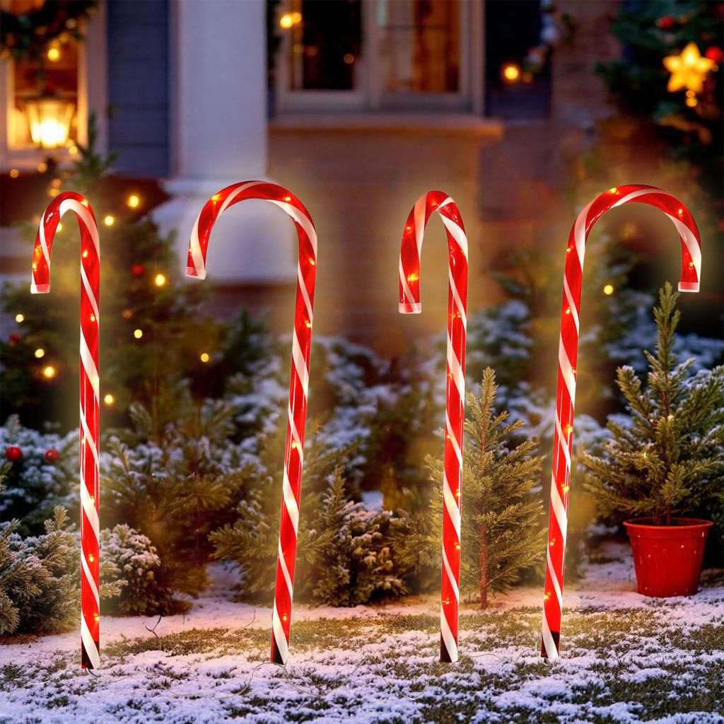 Four illuminated candy canes are placed in the snow in front of a house with Christmas lights.