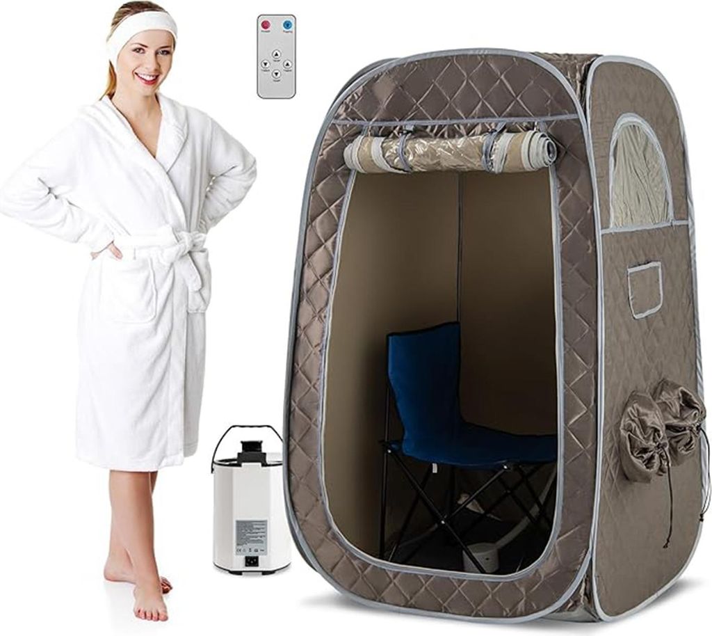 A woman in a white robe stands next to a portable steam sauna tent, a steam generator, and a remote control.