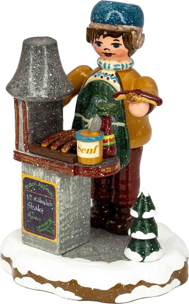 Ein fröhlicher Junge in winterlicher Kleidung steht an einem kleinen Bratwurststand mit einem Tablett voller Würstchen und einem Senfglas während ein kleiner Tannenbaum den verschneiten Boden schmückt