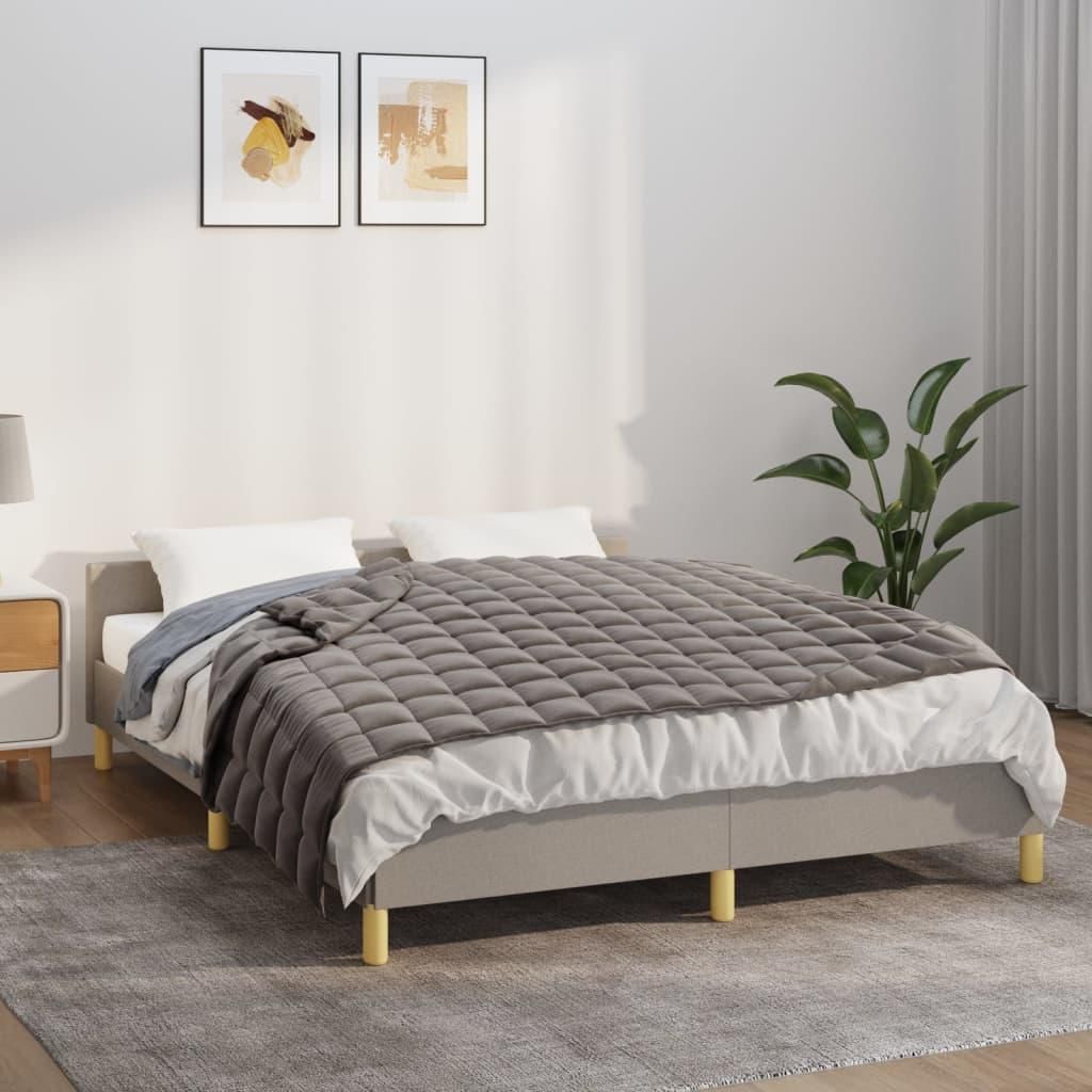 A bed with a gray weighted blanket and white bedding, set in a bedroom with artwork on the wall and a plant.