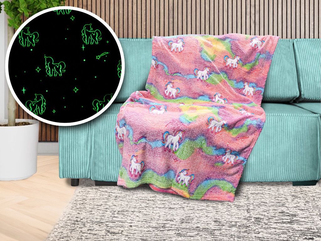 A fluffy blanket with a unicorn and rainbow pattern is draped over a teal sofa. A circular inset shows a close-up of the blanket's glow-in-the-dark unicorn pattern on a black background.