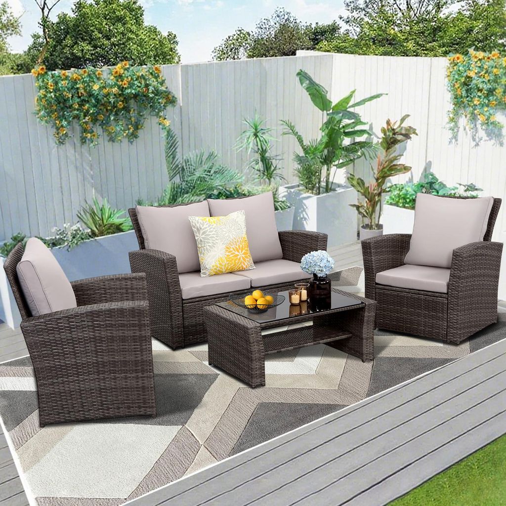 A patio furniture set is arranged on a rug on a wooden deck, with plants and a white fence in the background.
