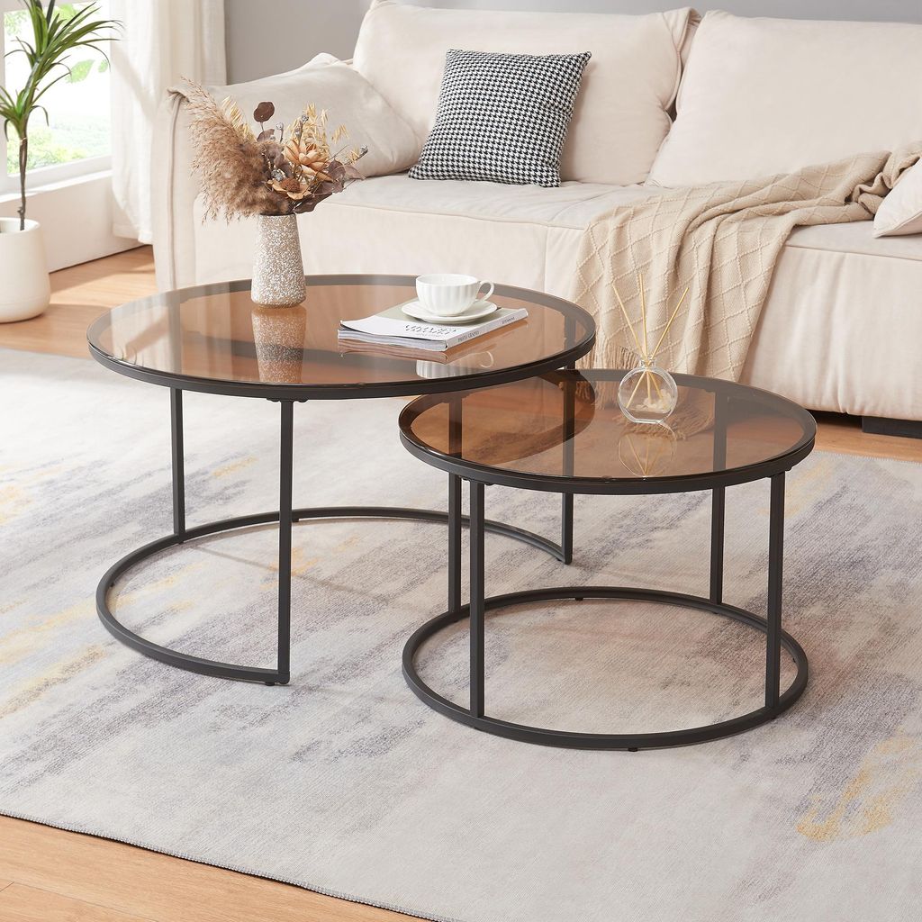 Two round coffee tables with brown glass tops and black metal frames are placed on a rug in front of a sofa.