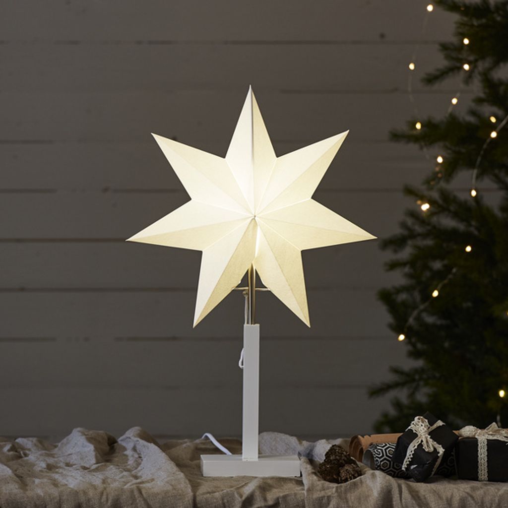 A lit paper star decoration on a stand, placed on a table with Christmas gifts and a decorated Christmas tree in the background.