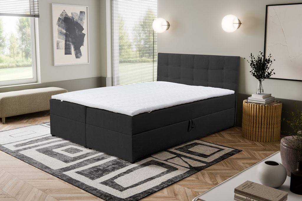 A bedroom scene featuring a black boxspring bed with a white mattress topper, a patterned rug, and decorative elements.