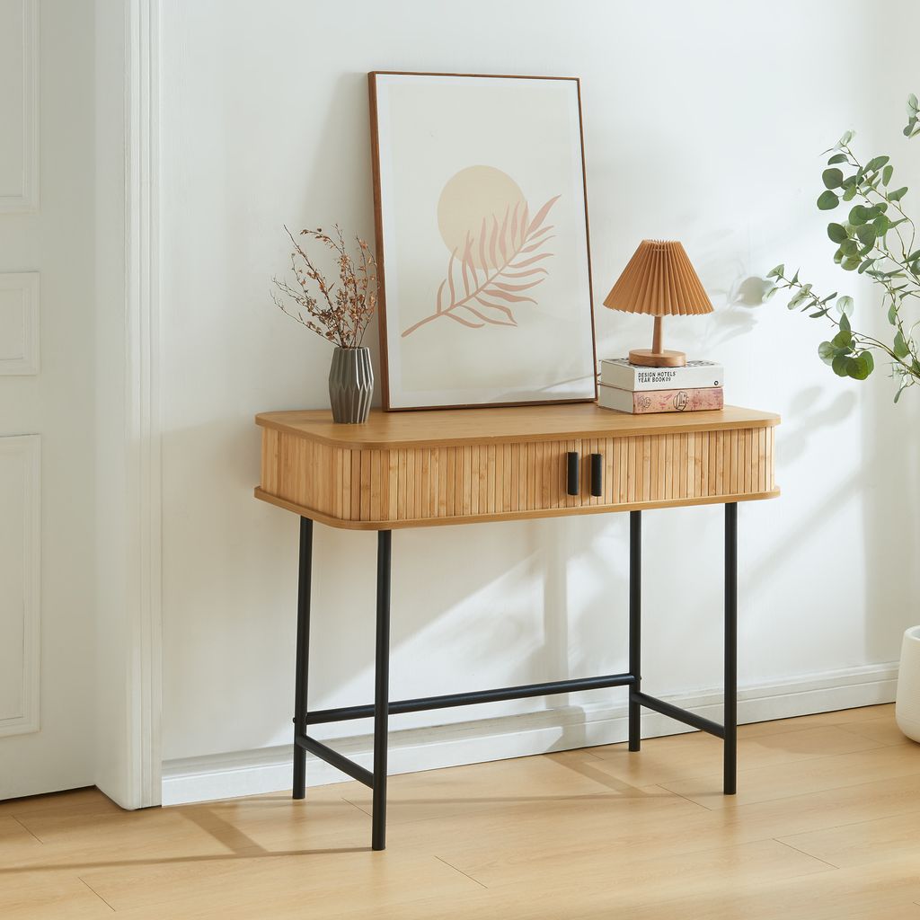 A console table with two doors and black legs, placed against a white wall with a framed picture and a lamp on top.