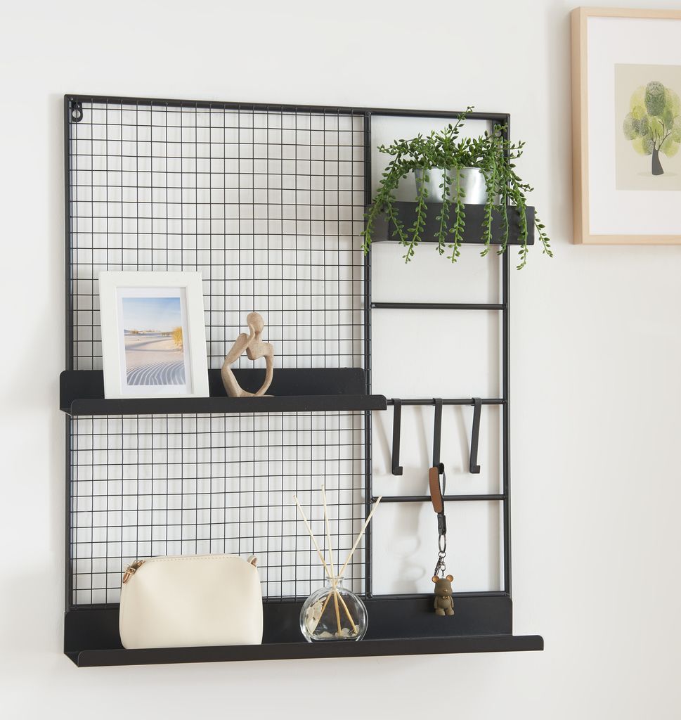 A black metal wall grid with shelves and hooks, decorated with a picture frame, a small sculpture, a plant, a purse, and a diffuser.