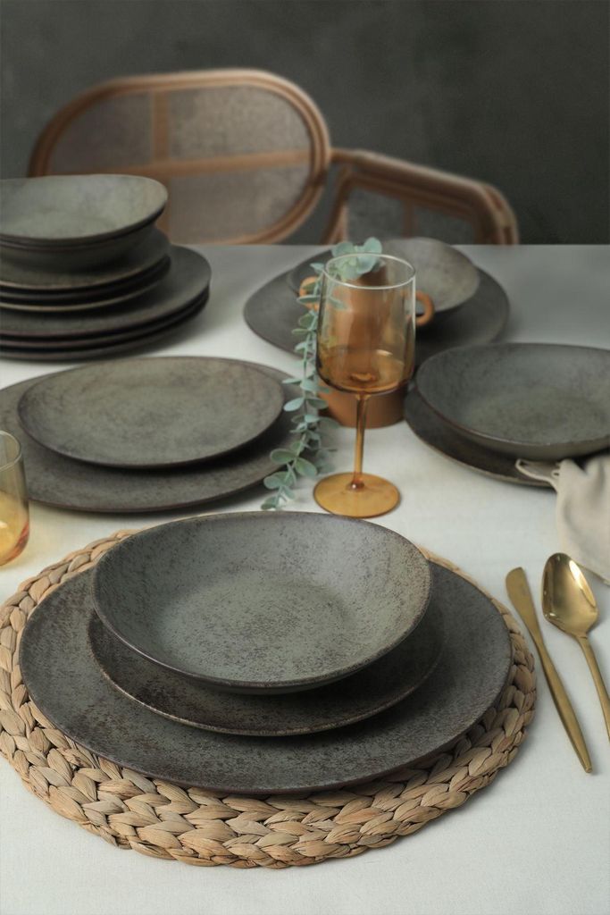 A table setting with stoneware plates, bowls, and a glass of amber liquid, with chairs in the background.