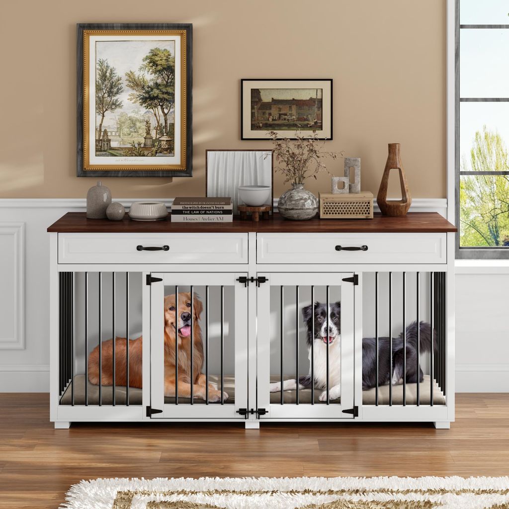 Two dogs are inside a white and brown dog crate furniture piece, which is placed on a wooden floor in front of a wall with artwork and decorative items.