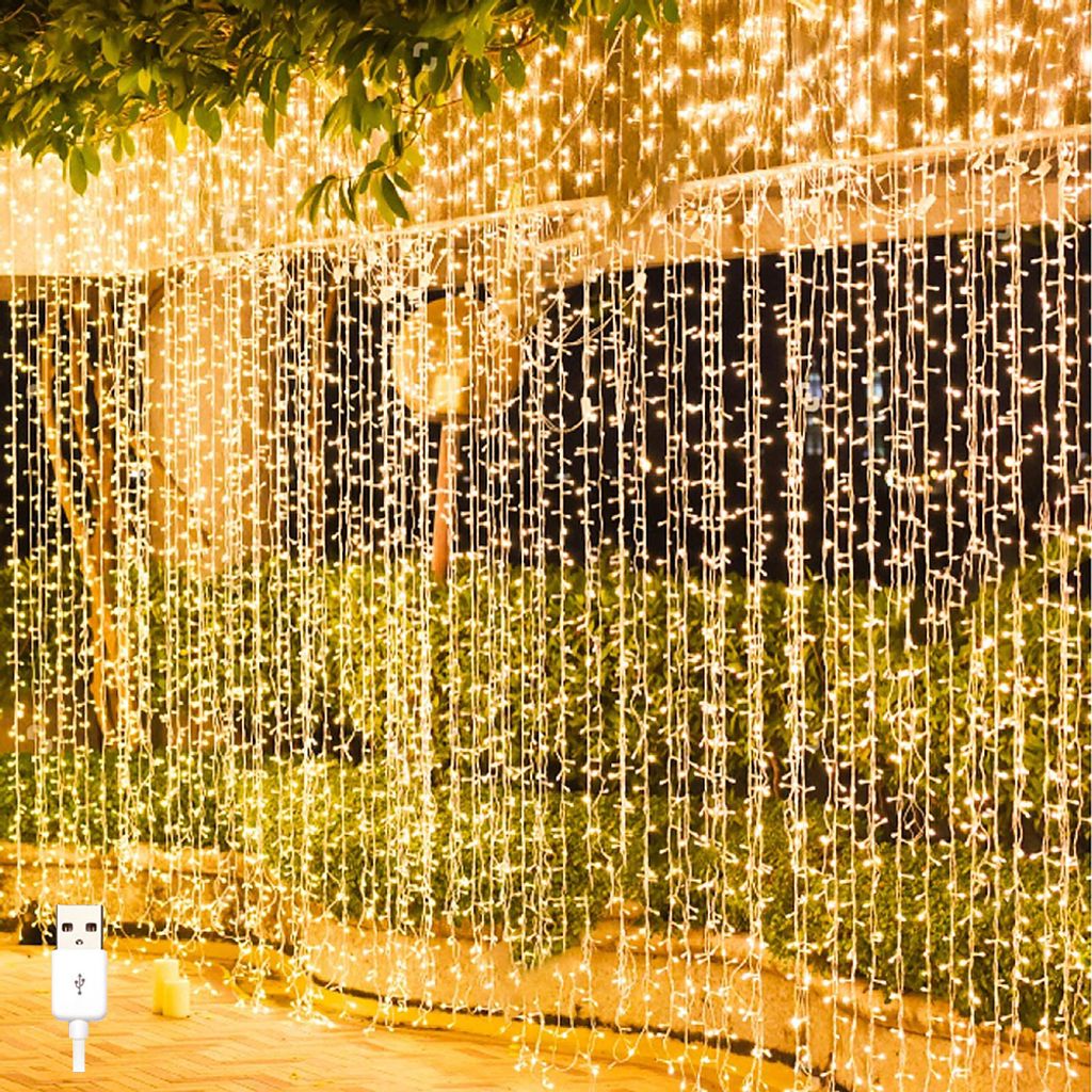 A curtain of warm white LED string lights hanging outdoors, possibly on a balcony or patio, with greenery in the background and a USB connector visible in the foreground.