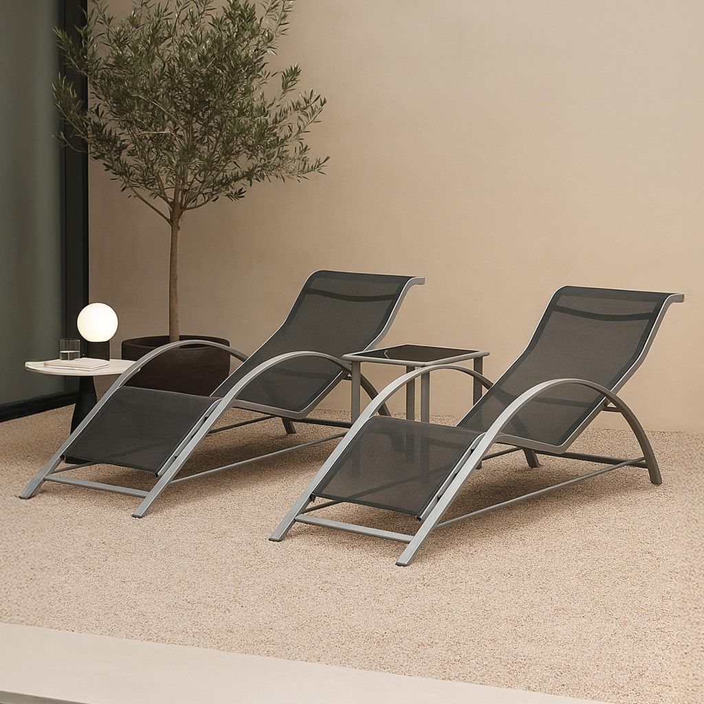 Two lounge chairs and a small table are set up on a textured outdoor surface, with a small tree and a wall in the background.