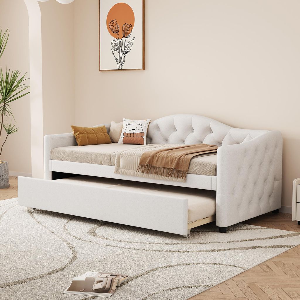 A white daybed with a pull-out trundle bed is shown in a room with a plant and artwork on the wall.