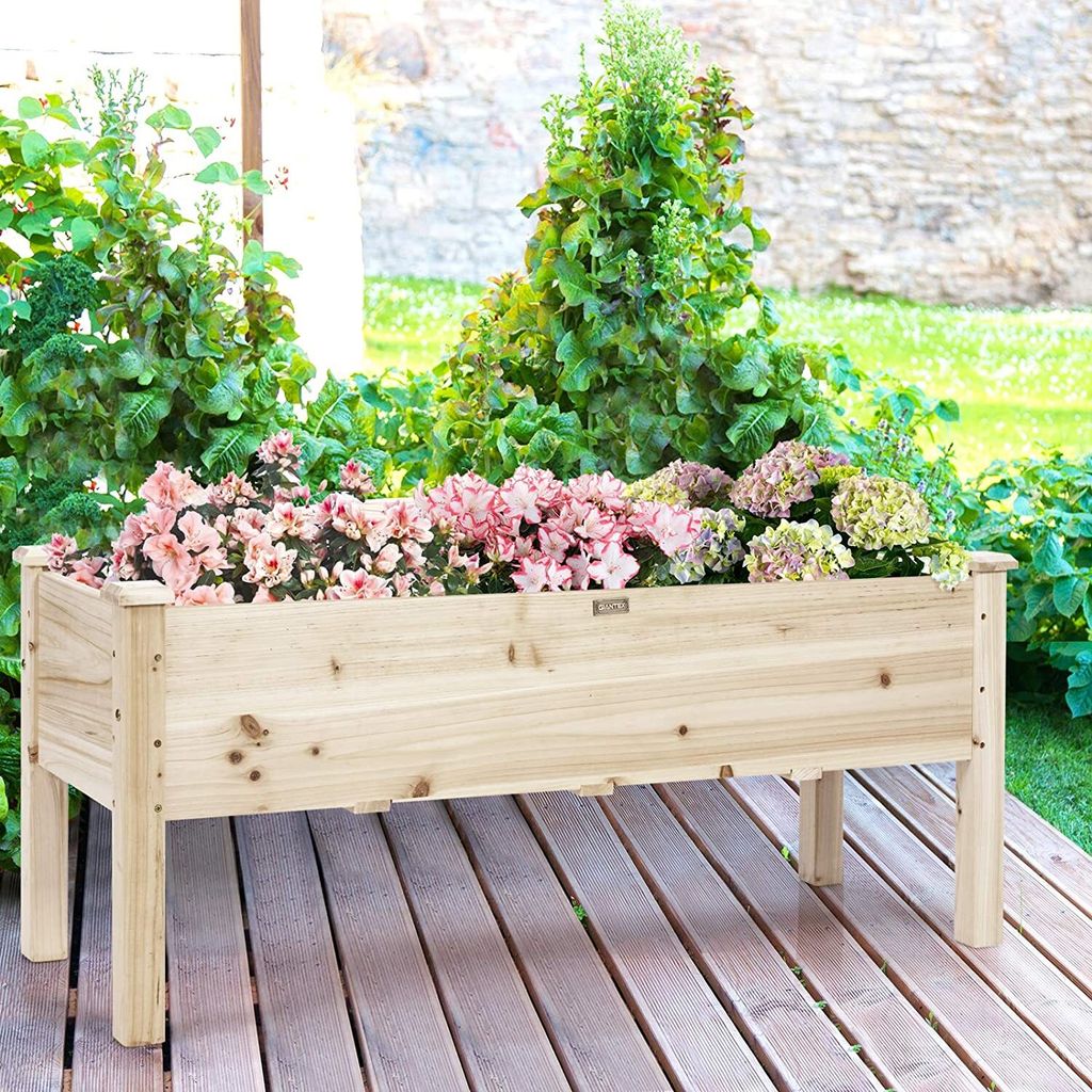 A wooden raised garden bed filled with pink and green flowers and plants, situated on a wooden deck outdoors.