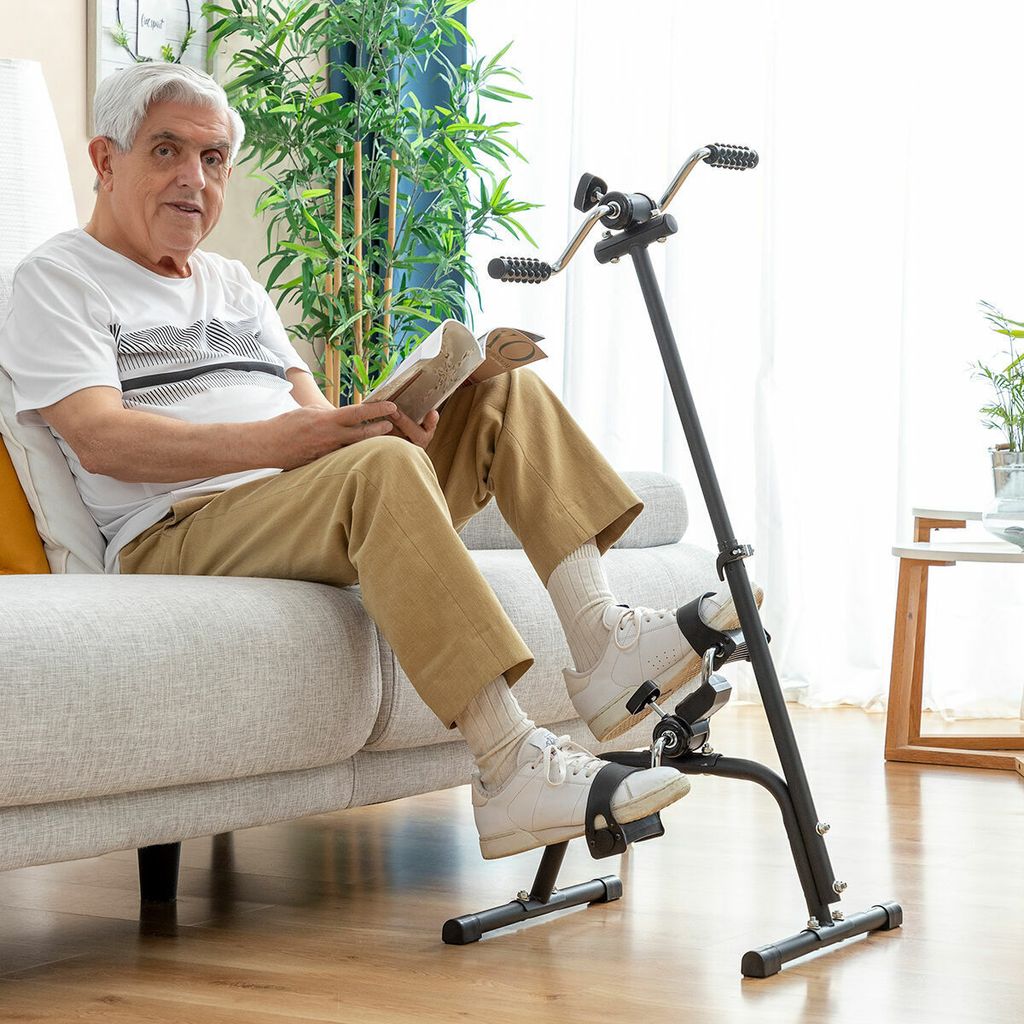 An elderly man sits on a couch reading a book while using a pedal exerciser for his legs.