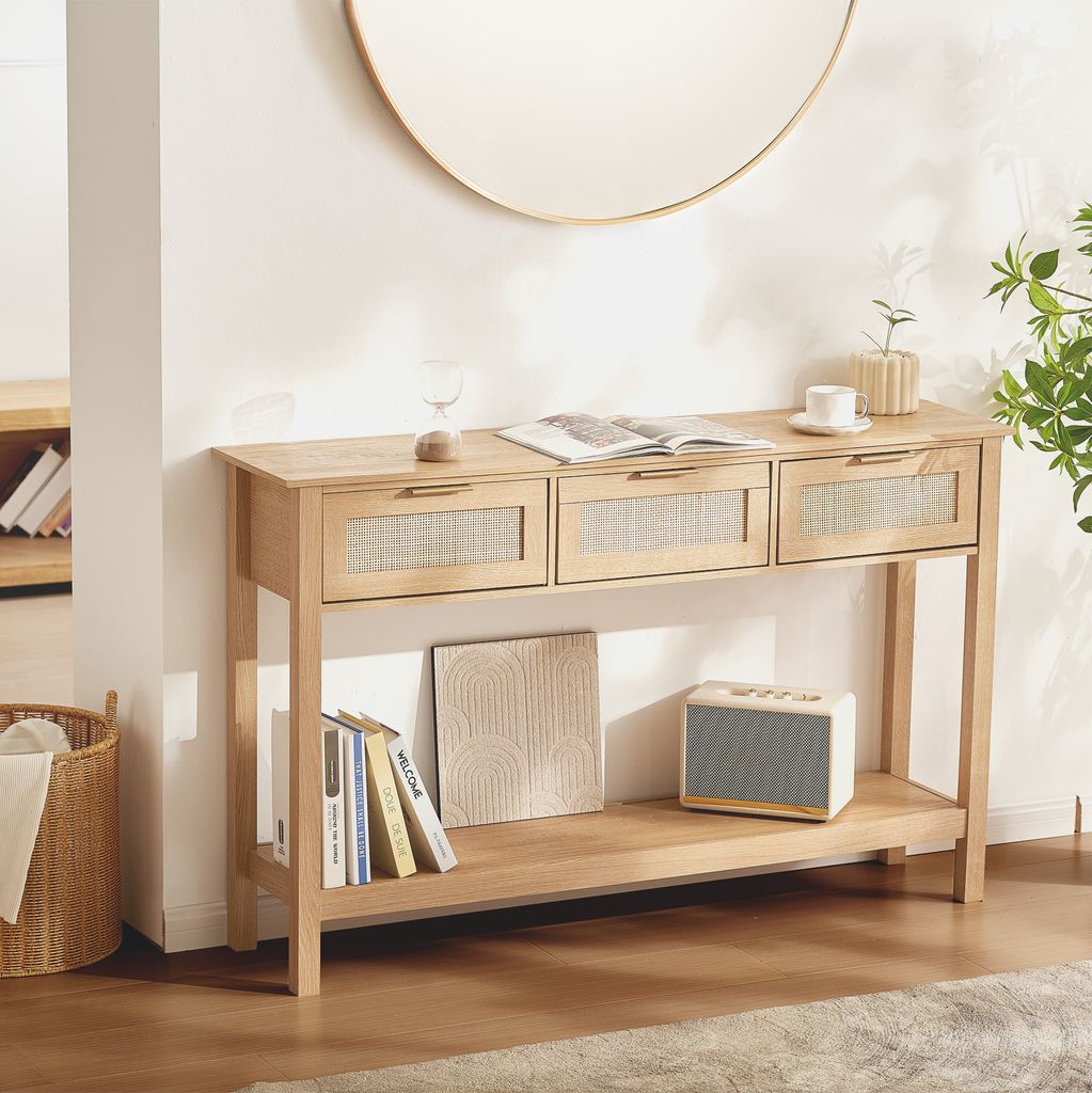 A light wood console table with three rattan-front drawers is placed against a white wall, reflecting a warm, inviting atmosphere. A large round mirror hangs above, and decorative items like books, a speaker, and a plant add to the scene.