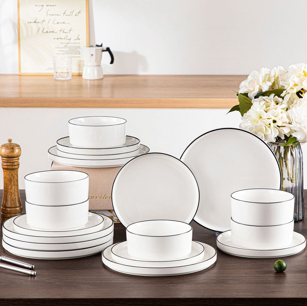 A set of white ceramic dishes with black trim is arranged on a wooden table, with a vase of flowers and a coffee maker in the background.