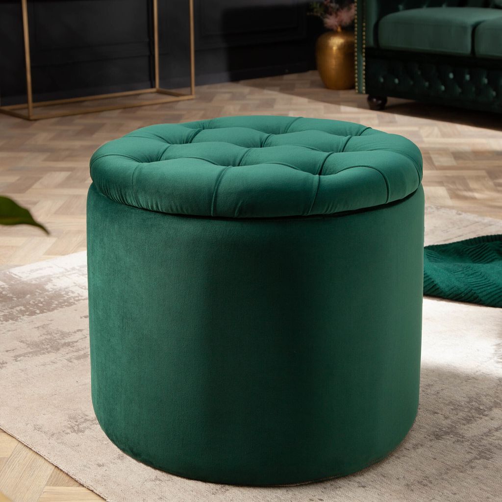 A dark green velvet ottoman with tufted top sits on a rug in a room.
