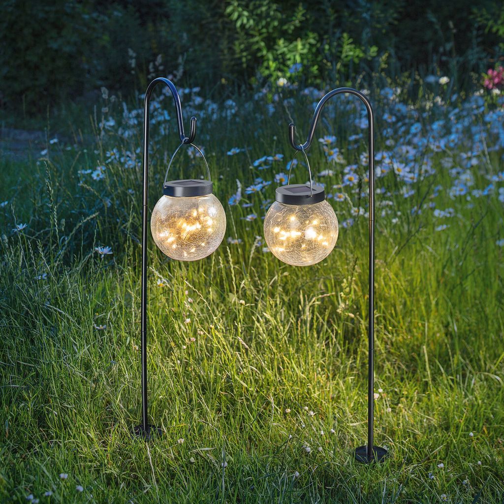 Two solar-powered globe lights hanging from hooks in a grassy outdoor setting with wildflowers.