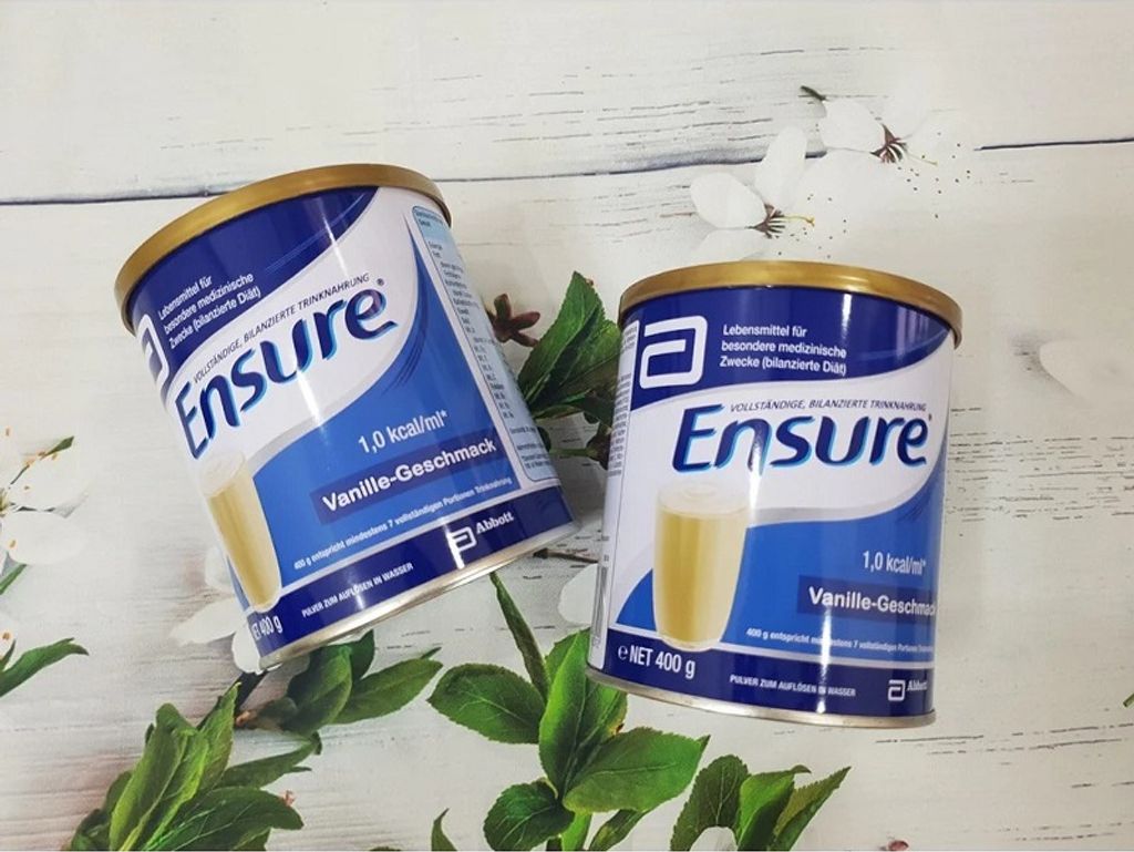 Two cans of Ensure vanilla flavored powder drink mix are displayed on a wooden surface with green leaves and white flowers.