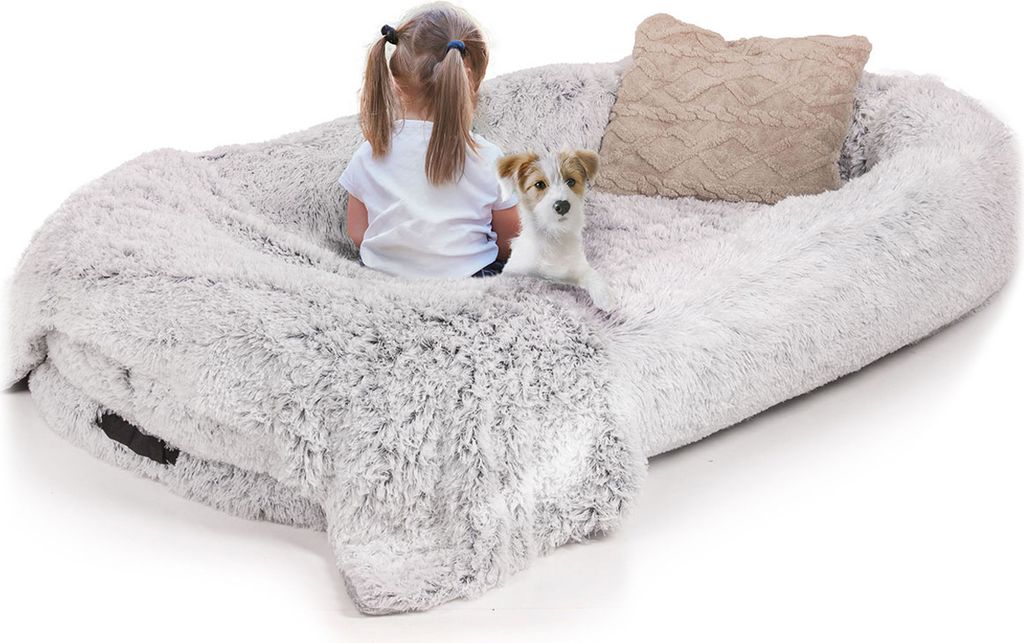 A young child and a small dog are sitting on a large, plush, light gray pet bed with a beige pillow.