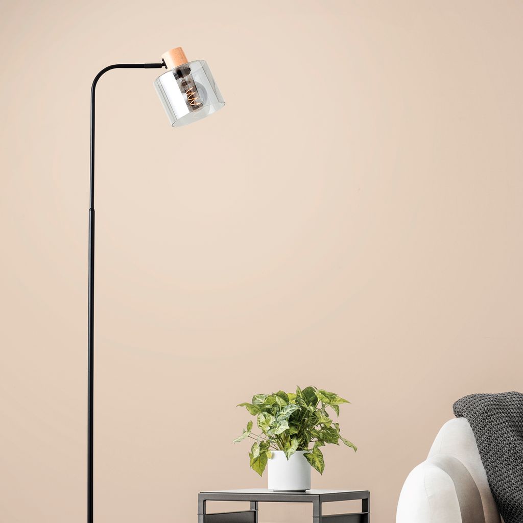 A modern floor lamp with a glass shade and wooden accents is placed next to a sofa with a blanket, with a small potted plant on a side table in the foreground.