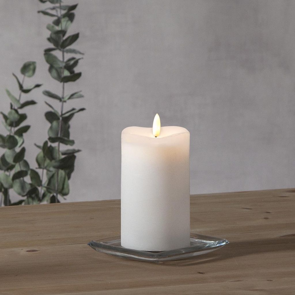 A white LED candle with a flickering flame sits on a square glass coaster on a wooden table. In the background, a grey wall is visible with a branch of eucalyptus leaves.