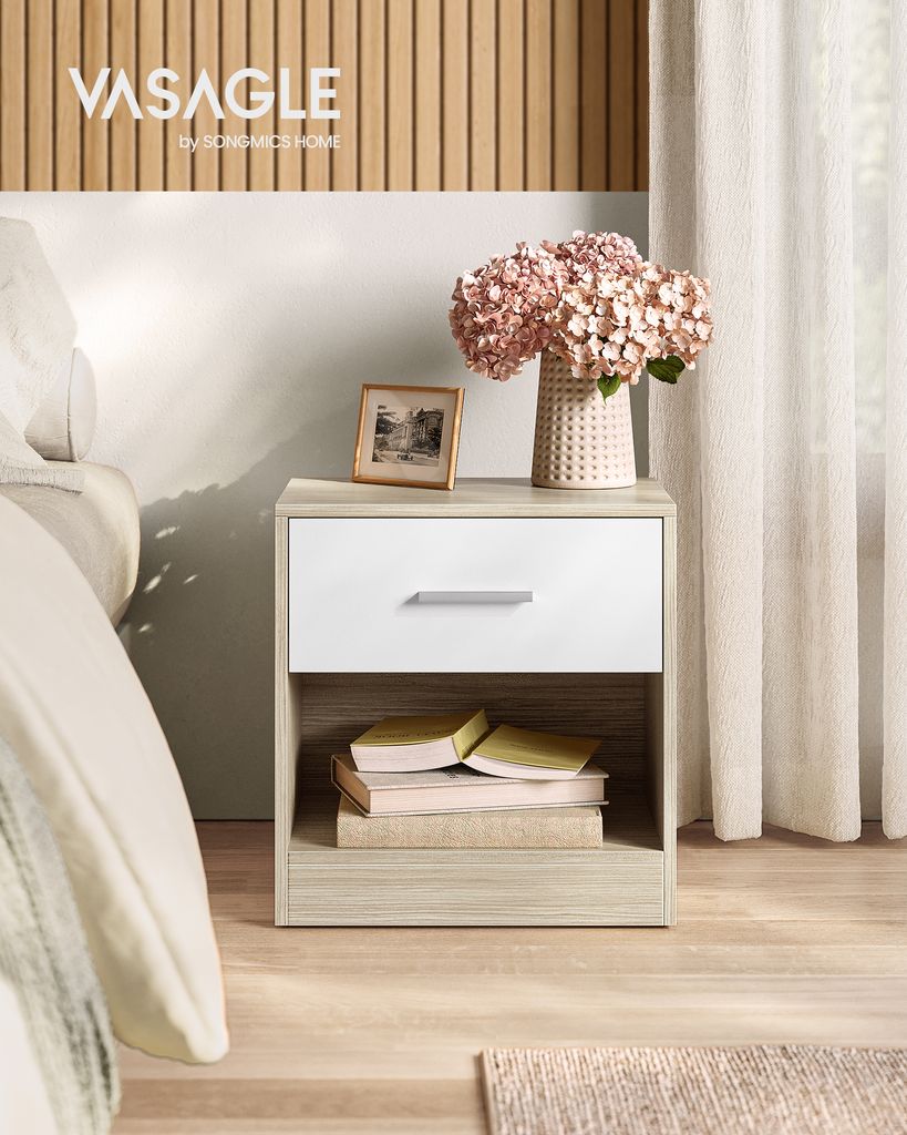 A nightstand with a drawer and open shelf next to a bed, decorated with flowers and a picture frame.