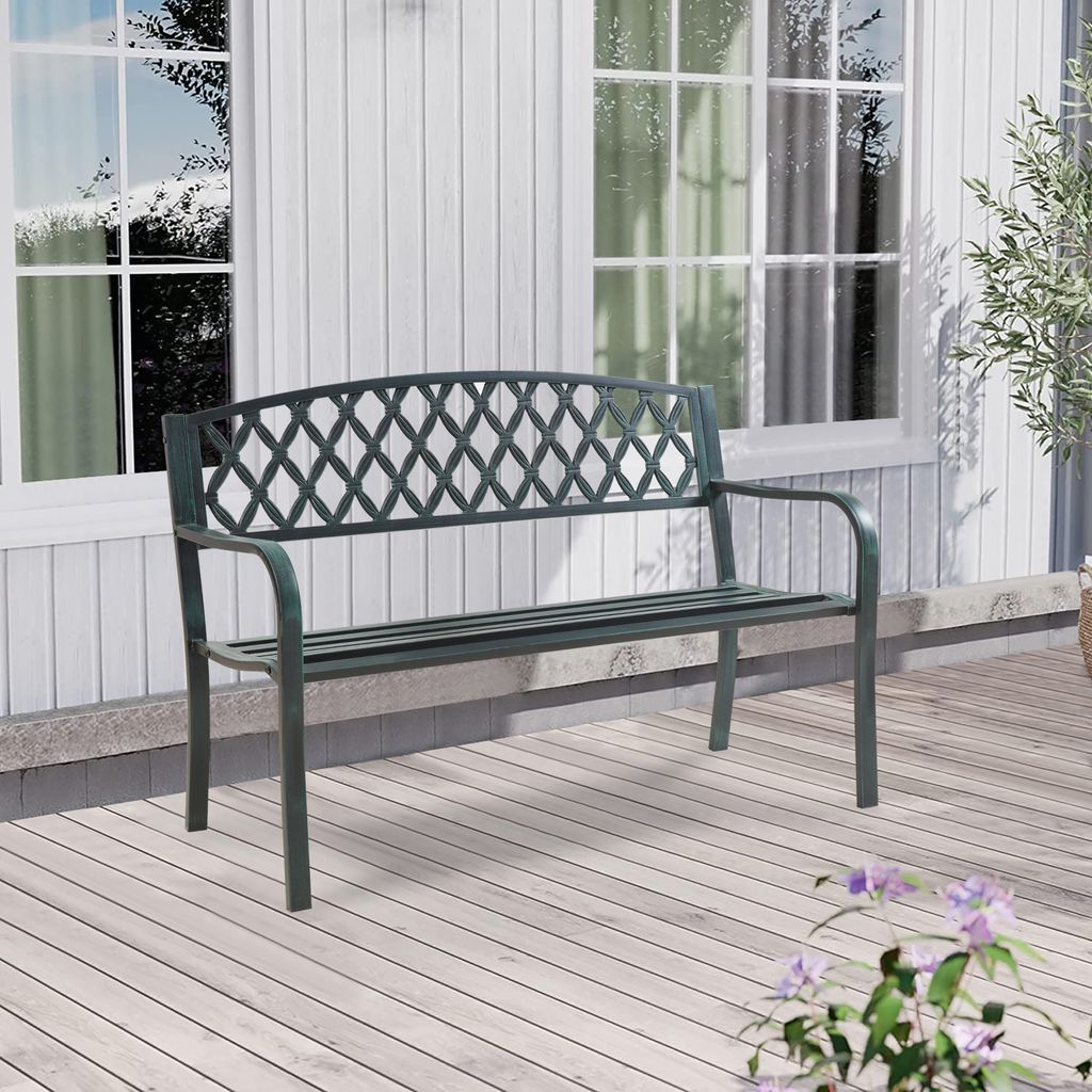 A dark gray metal garden bench with a green patina sits on a light-colored wooden deck in front of a white-paneled house with windows.