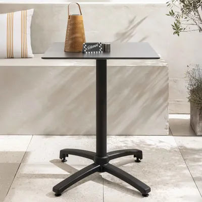 A square black outdoor table with a black base and a small sign on top, placed on a tiled patio next to a bench with a striped cushion and a woven lantern.